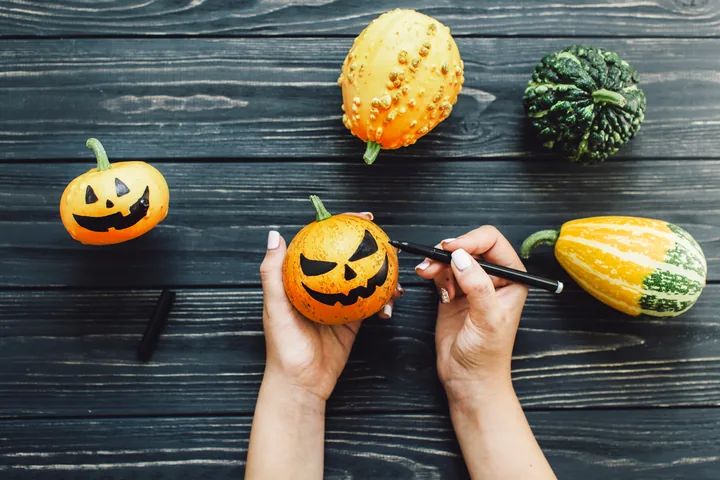 Hands draw a black face on a small pumpkin beside another decorated pumpkin and several gourds.