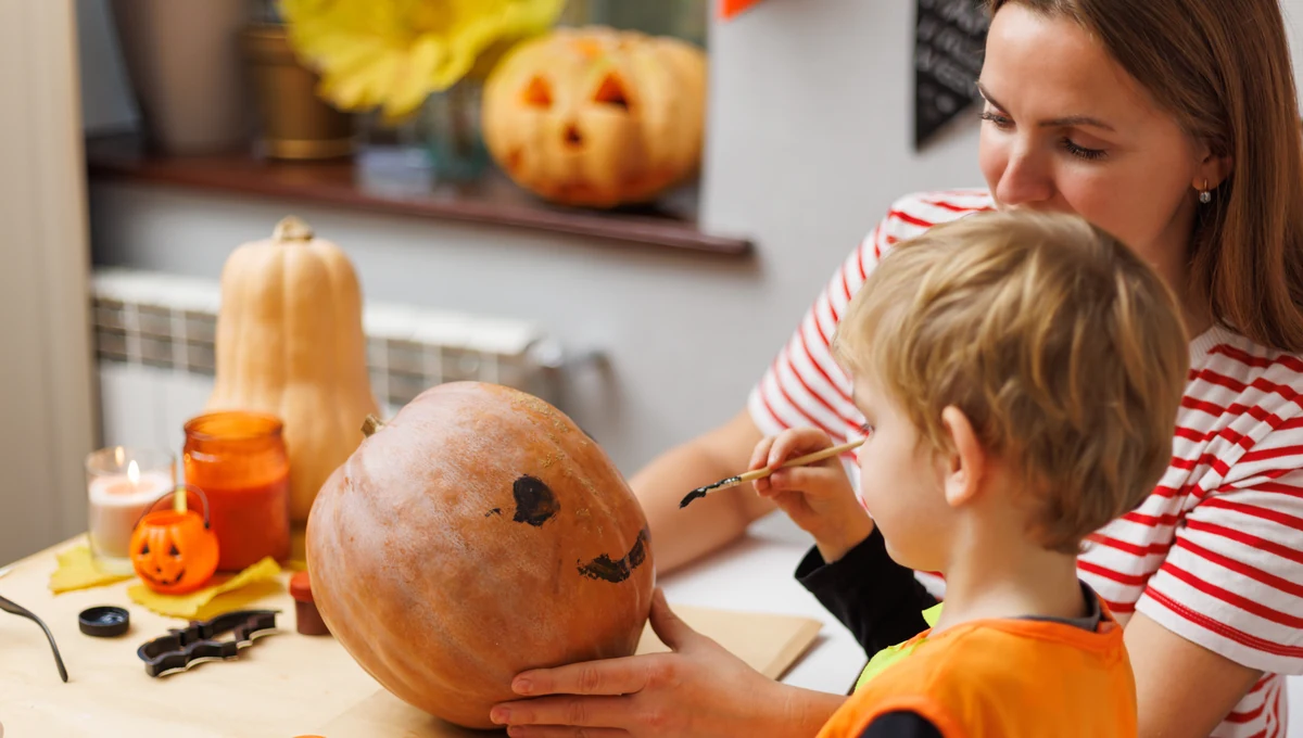 Halloween pumpkin painting with a parent A preschooler paints a pumpkin with a paintbrush while their mother holds it steady.