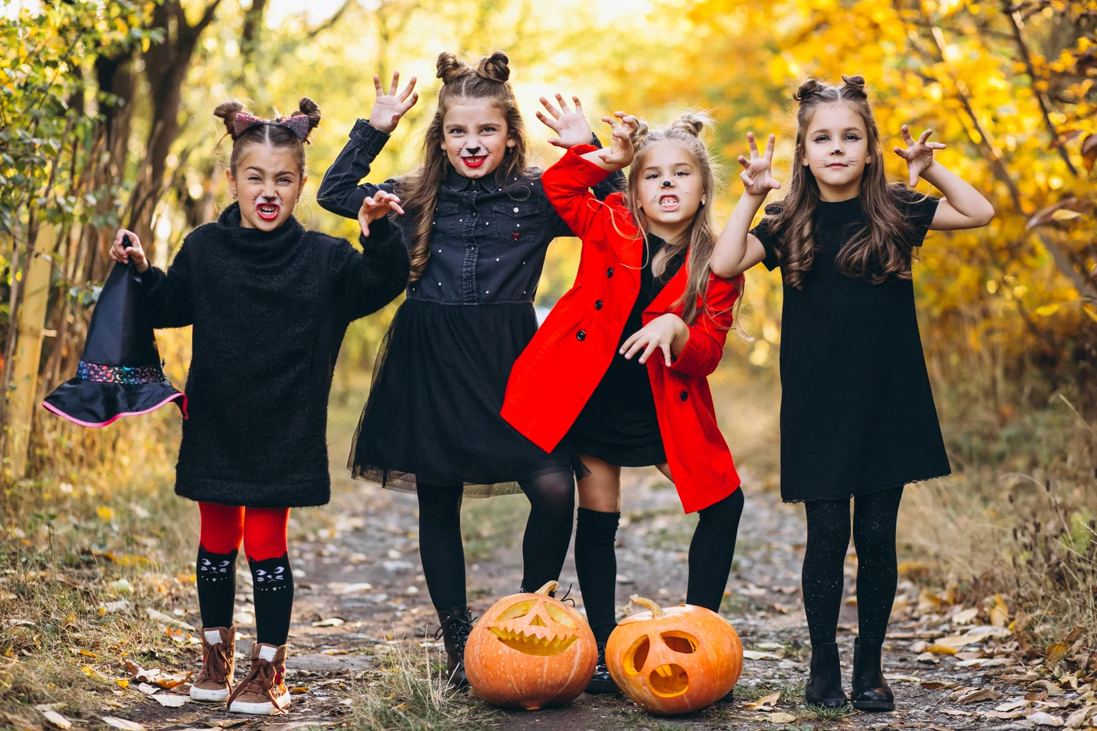 Four children in dark outfits and Halloween face paint playing outdoors among autumn leaves and pumpkins.