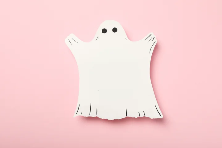 White paper ghost cutout with black eyes on a pink background.