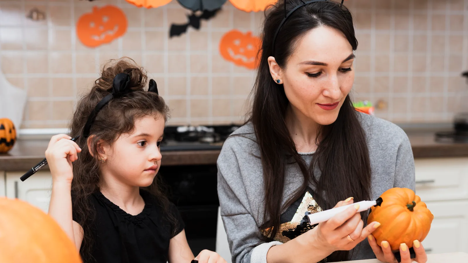 A mother draws on a small pumpkin while her daughter watches and holds a black marker.