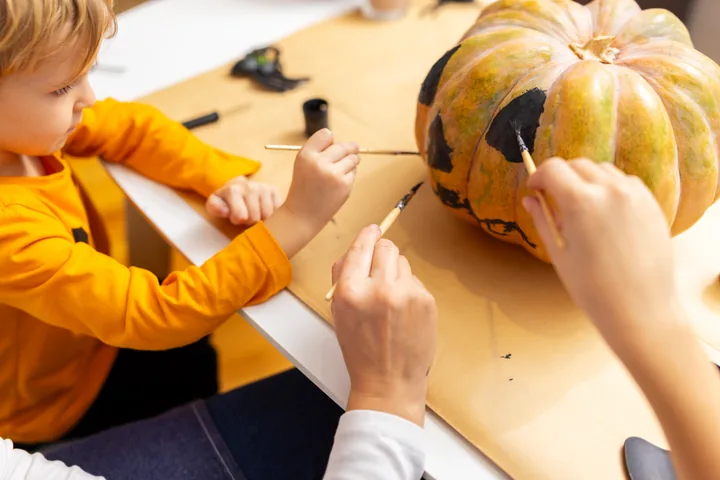 A child and grown-up use paintbrushes to paint black eyes and a stitched mouth on a pumpkin.